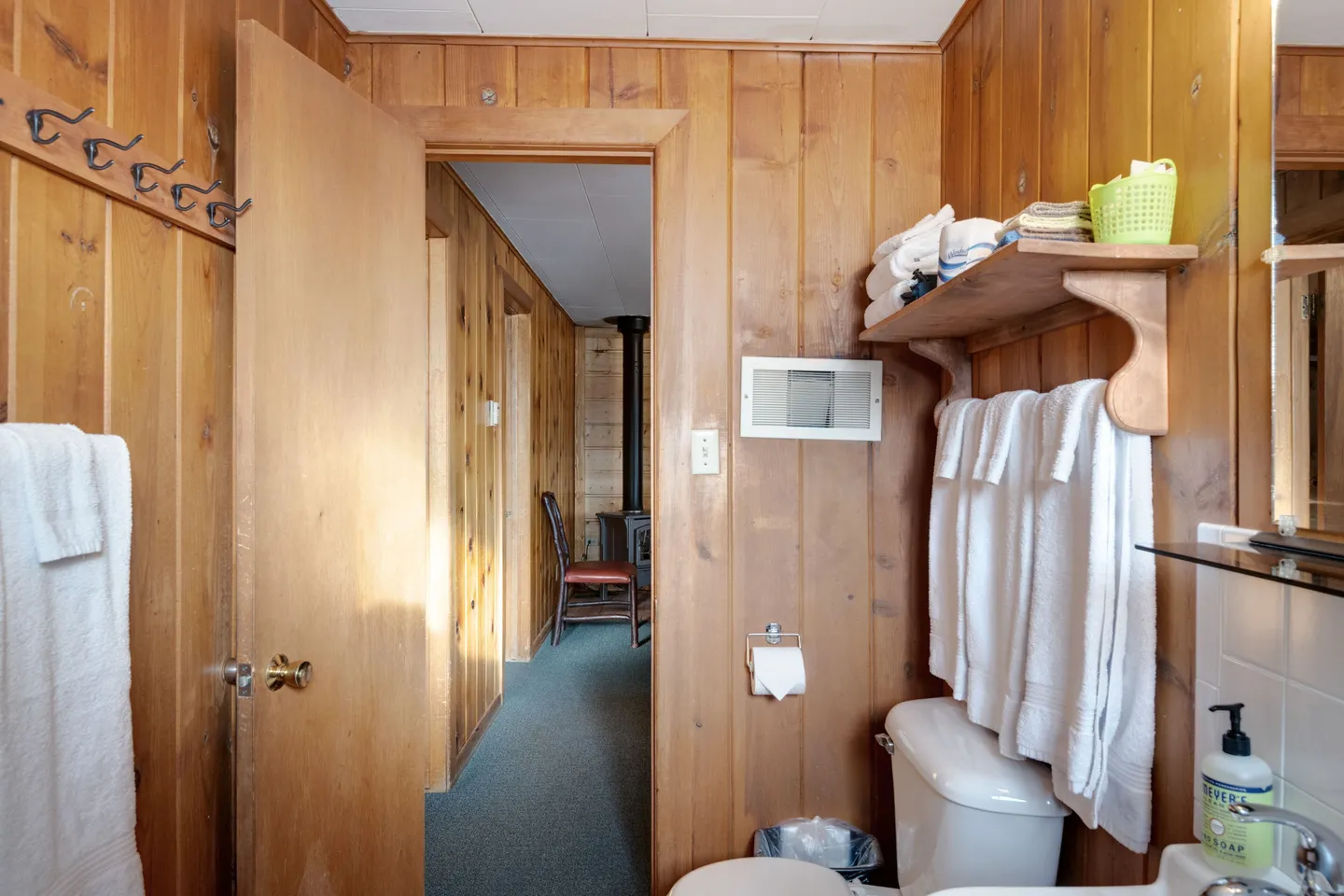 Bathroom with wood-paneled walls, an open doorway showing a carpeted hallway, toilet, and wooden shelf with white towels.