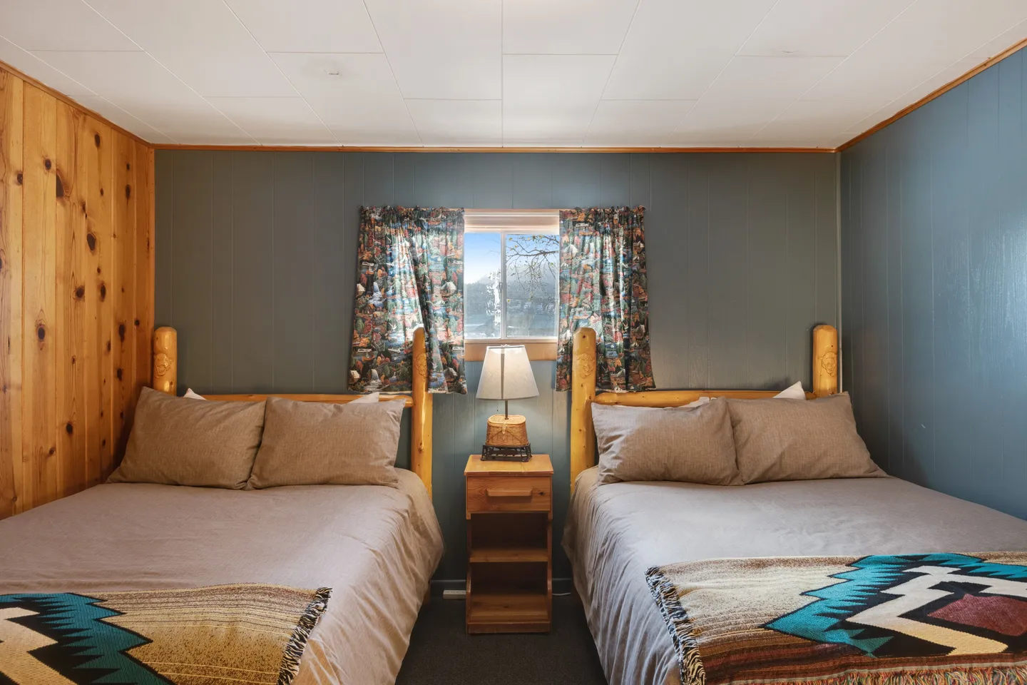 Bedroom with two log-style beds separated by a small nightstand with a lamp under a window.