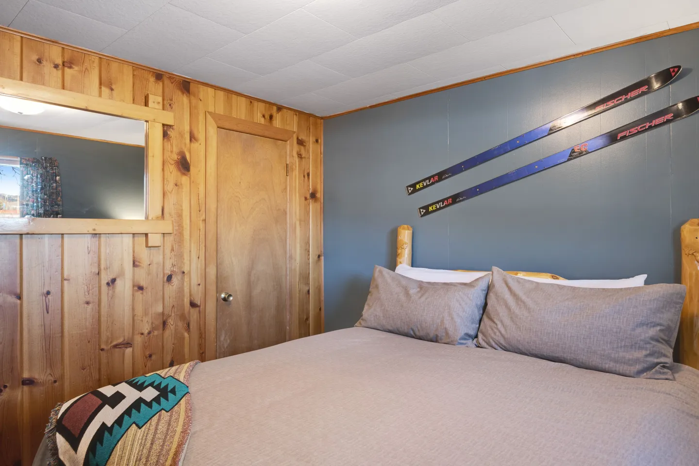 Bedroom with wood-paneled wall, blue accent wall, pair of decorative skis, and made bed with log-style headboard.