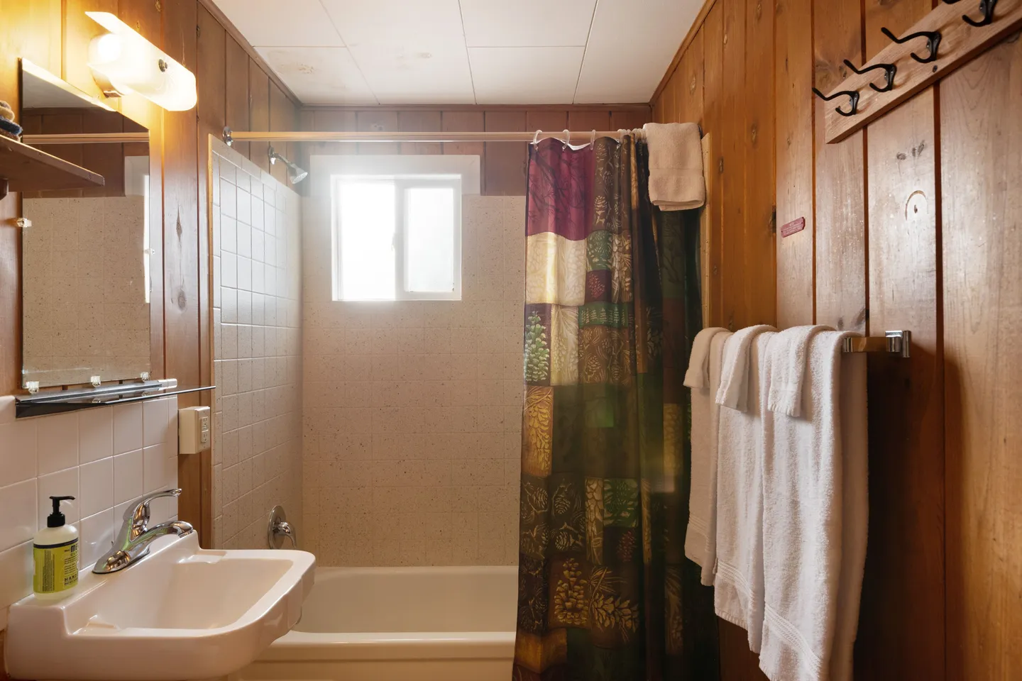 Bathroom with wood-paneled walls, a white sink, mirror with vanity light, and a bathtub with shower curtain and small frosted window.