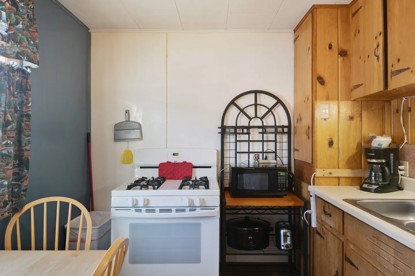 Kitchen corner with white gas stove, a metal bakers rack holding a microwave and small appliances, and wooden knotty pine cabinets.