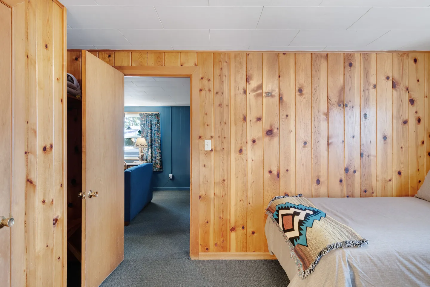Bedroom with knotty pine wood-paneled walls, carpeted flooring, a made bed, and an open doorway looking into a living room with blue walls.