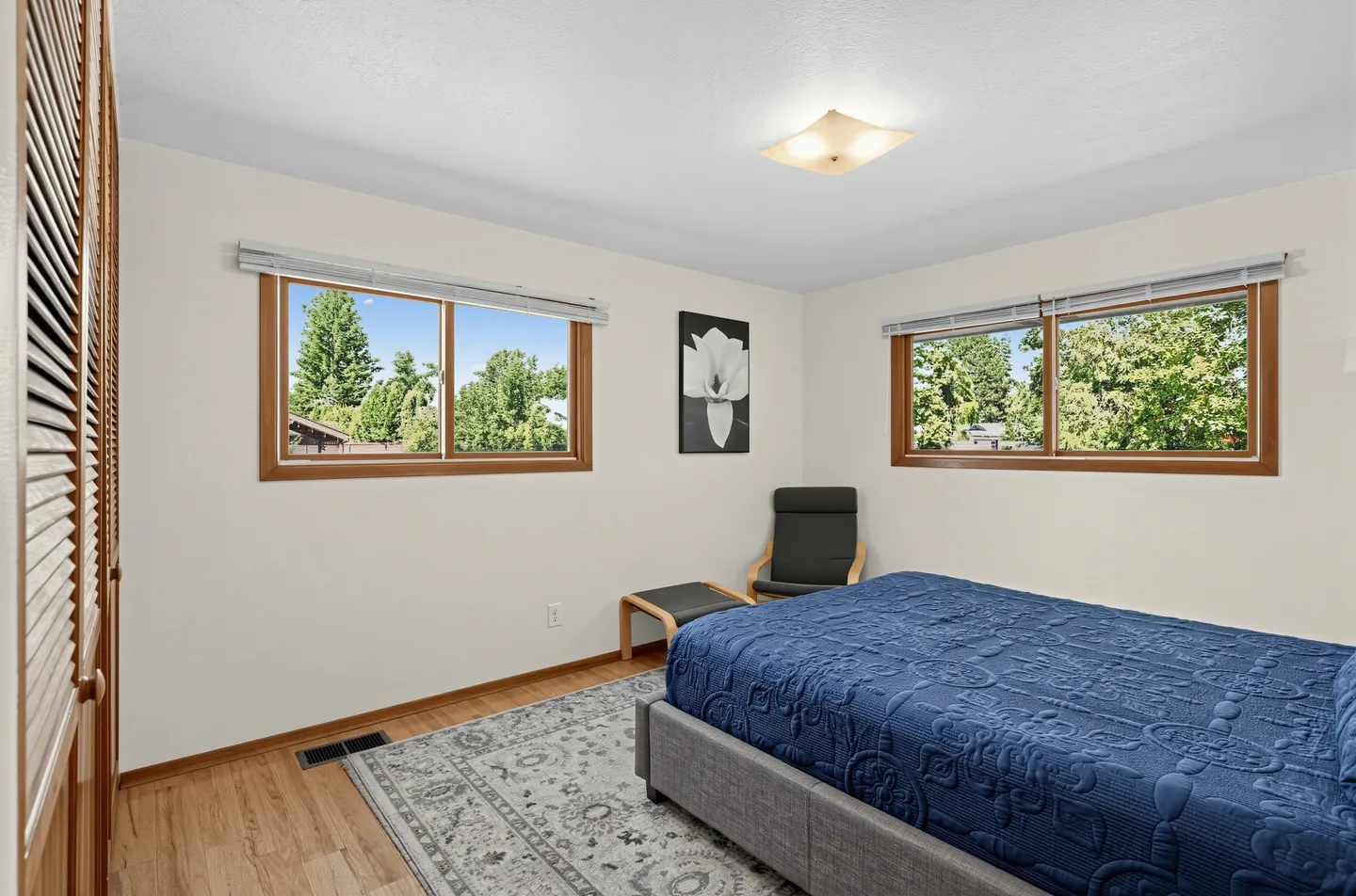 Bedroom with a blue-covered bed, wood-framed windows overlooking trees, an armchair with an ottoman, and a pattern rug over wood-look flooring.