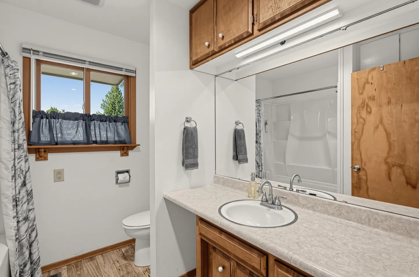Bathroom featuring a vanity with a large mirror, wood cabinetry, a toilet, and a window with blue curtains.
