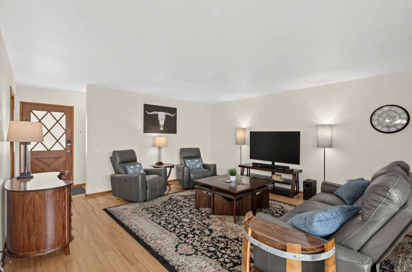 Living room with light wood floors, an ornate area rug, gray reclining leather seating, and a flat-screen TV.