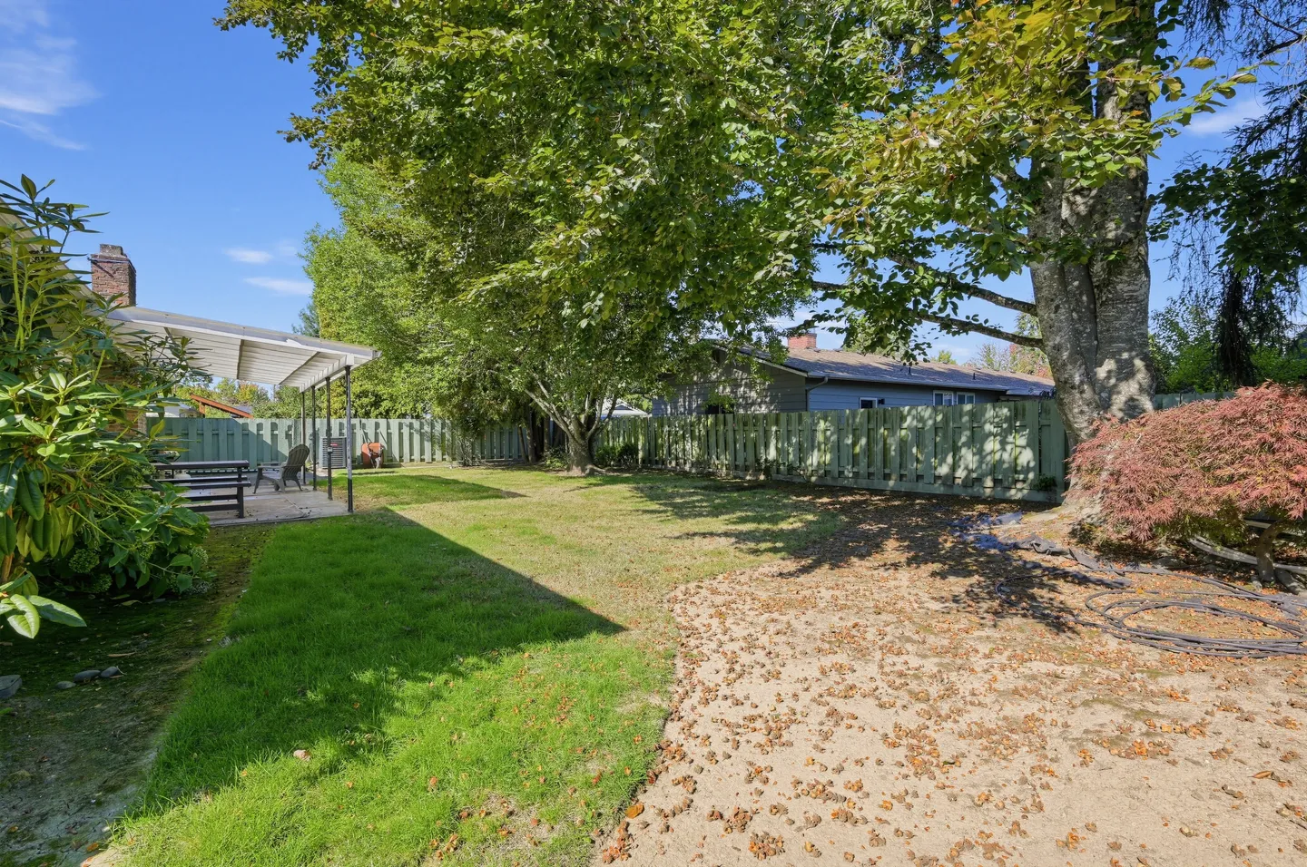 A fenced backyard featuring a grass lawn, large shade trees, a covered patio on the left, and a wooden perimeter fence.