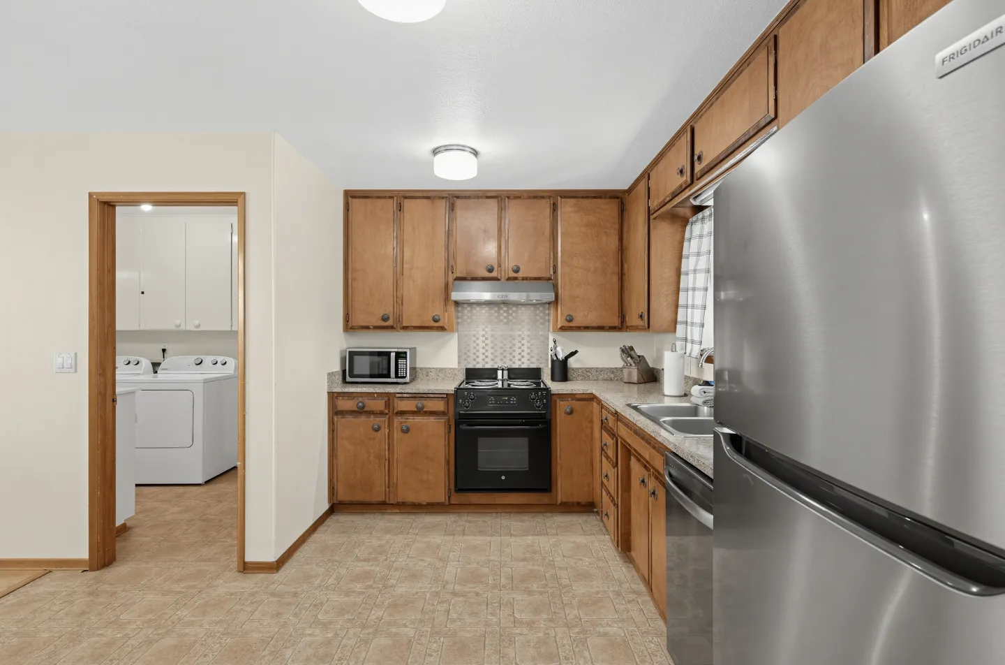 Kitchen with wooden cabinets, laminate countertops, a black stove, stainless steel refrigerator, and a doorway showing a laundry room.