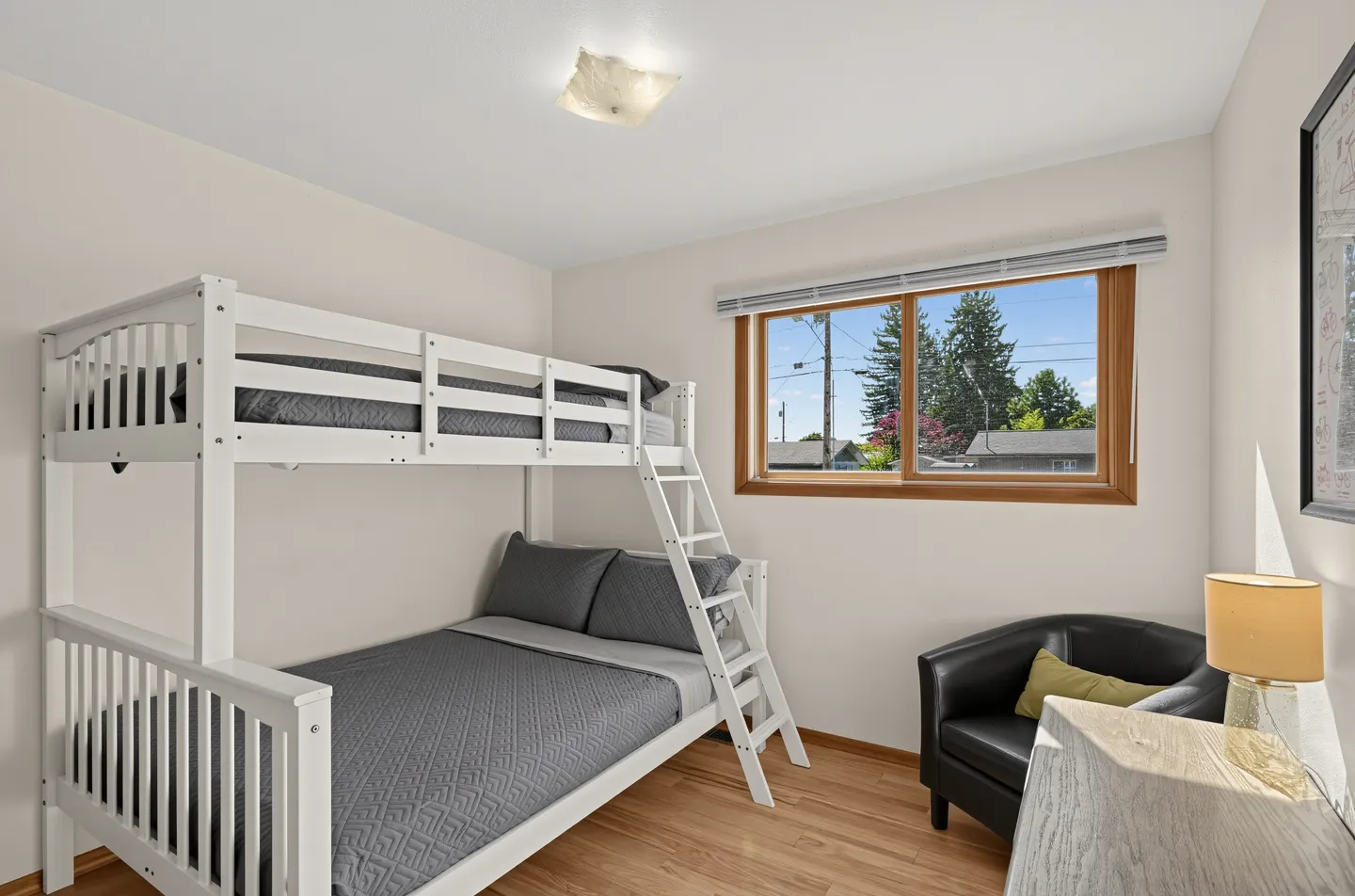 Bedroom featuring a white wooden bunk bed with grey bedding, a window with outdoor views, and a black armchair in the corner.