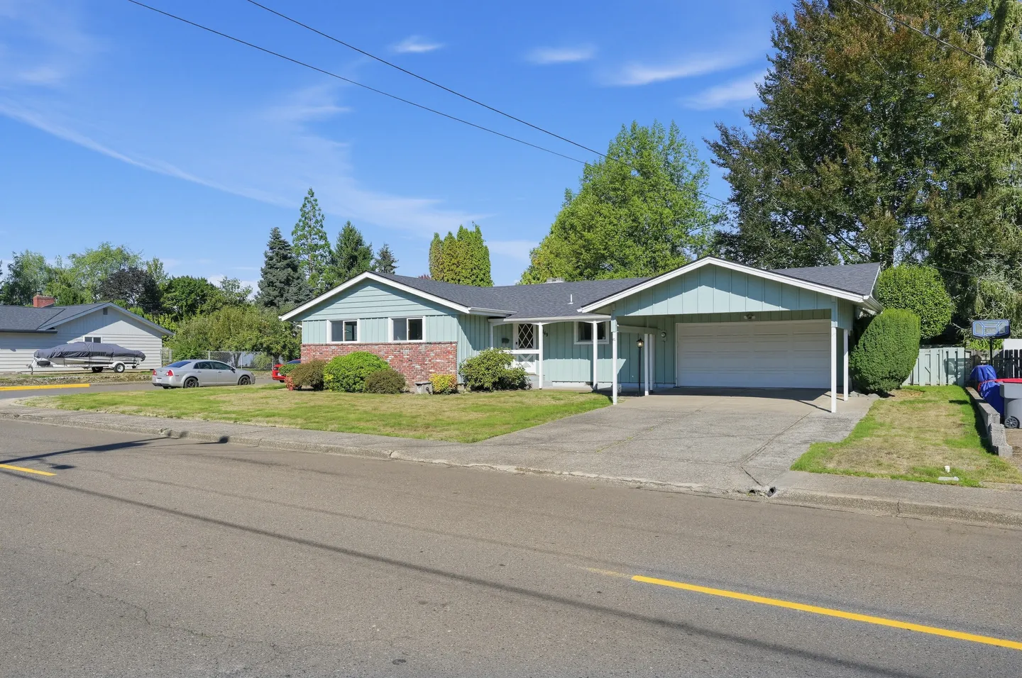 Street view of a single-story light green home with a driveway, carport, and front lawn under a clear blue sky.