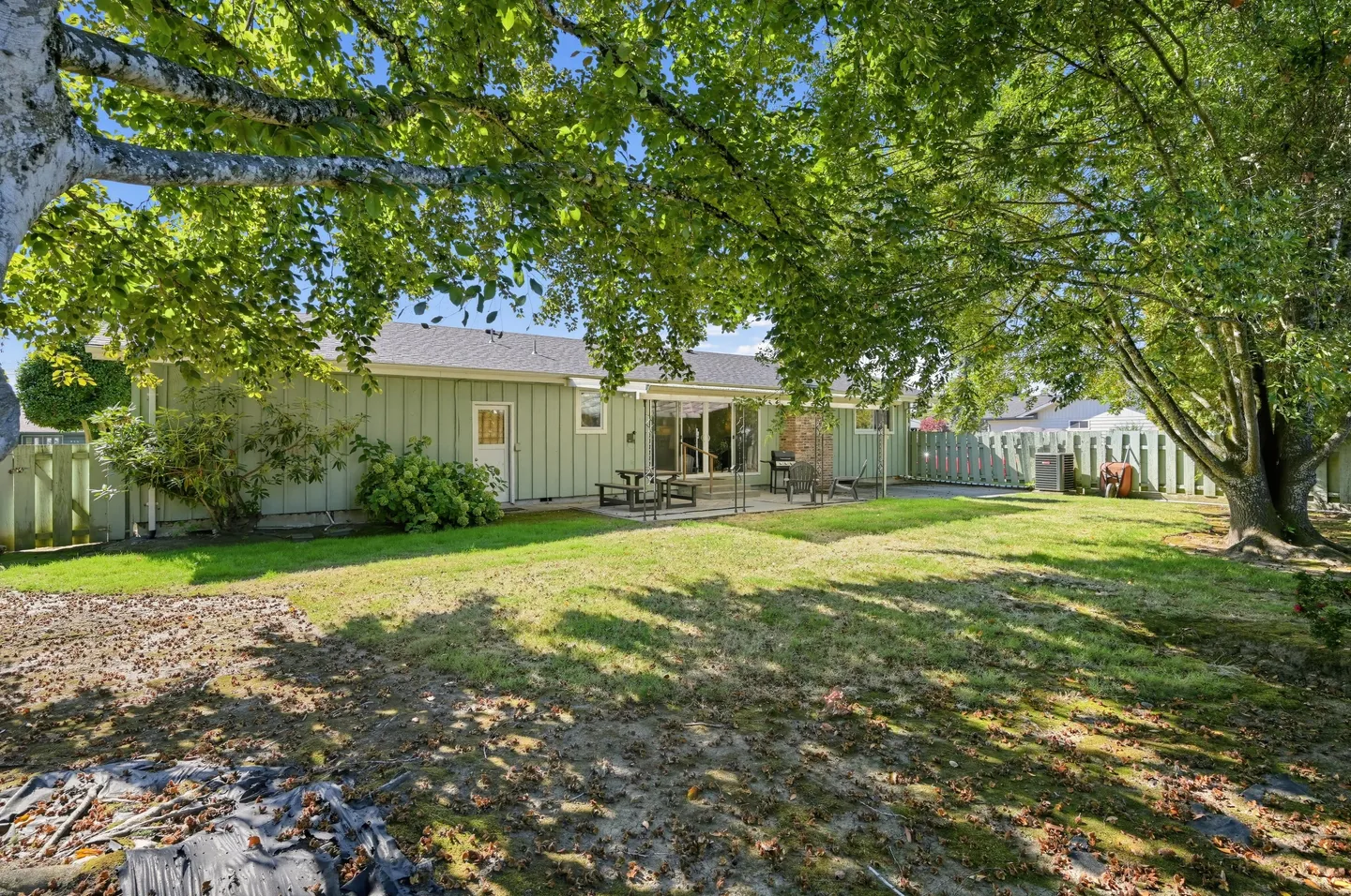 Fenced backyard and rear exterior of a single-story house with patio seating beneath mature shade trees.