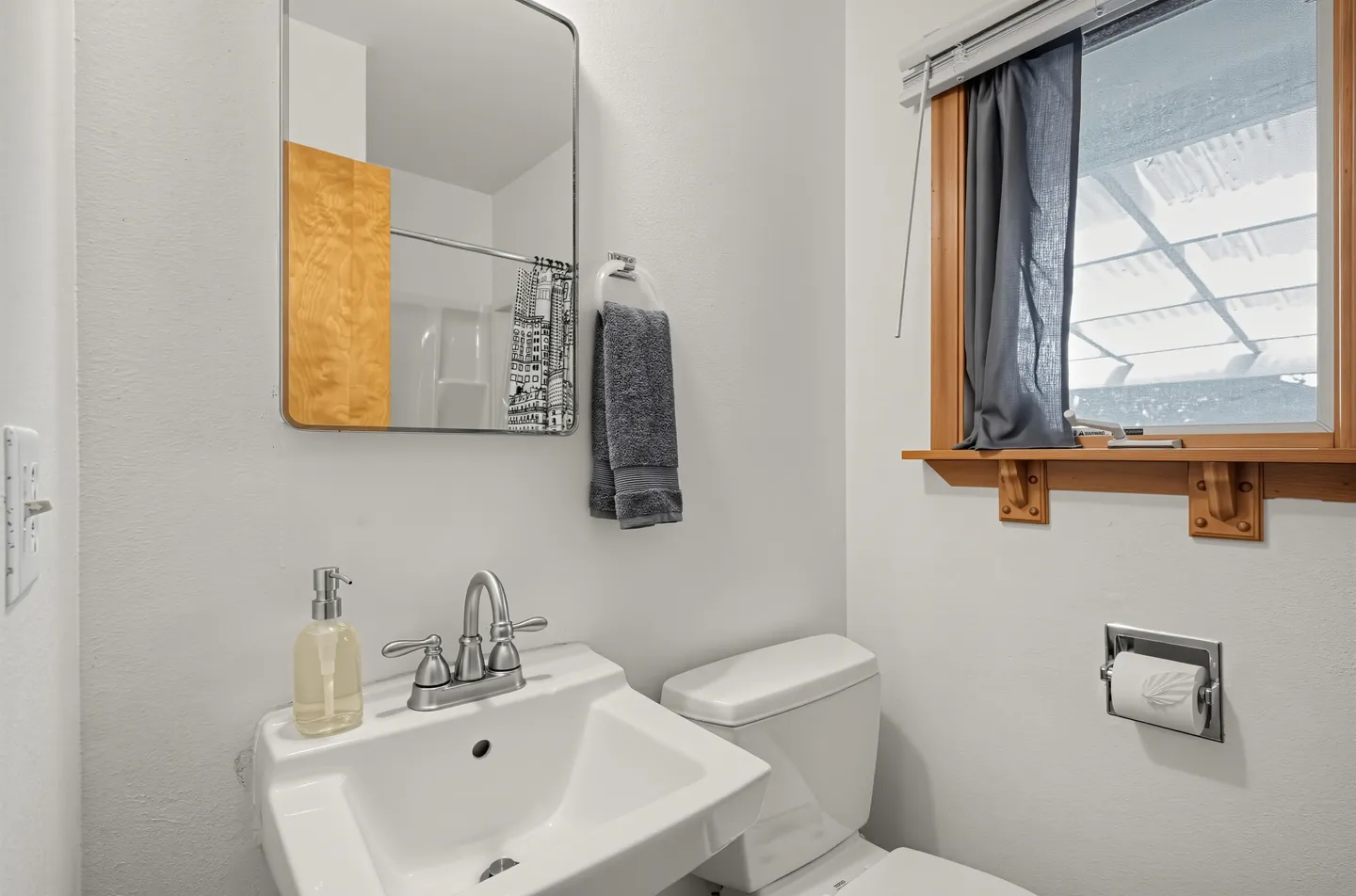 Bathroom with white pedestal-style sink, mirror, toilet, and window with a dark blue curtain.
