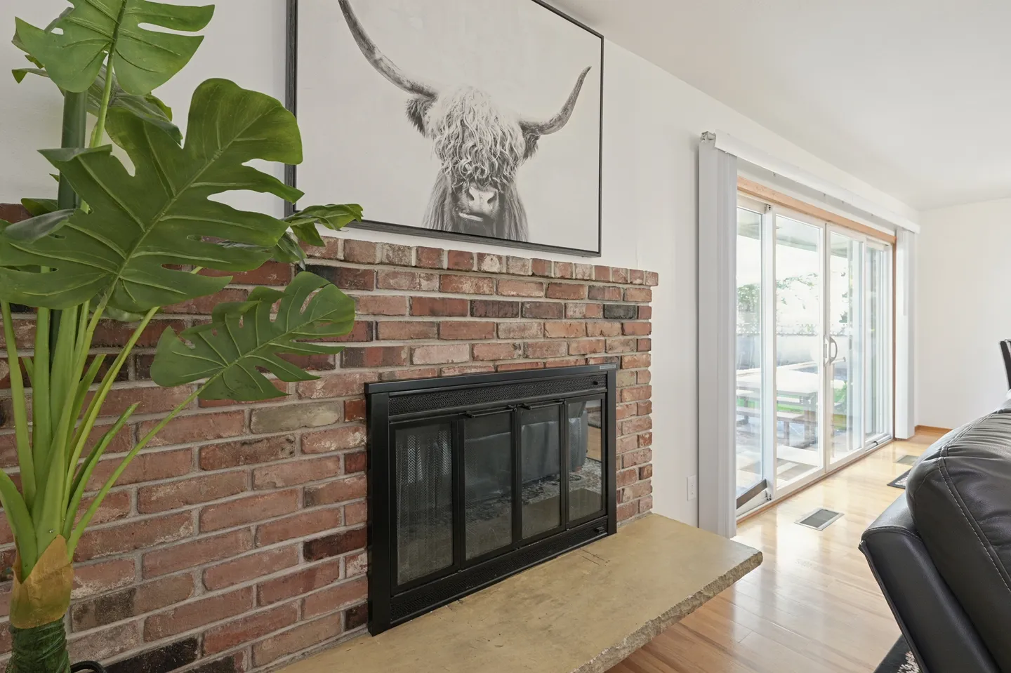 Brick fireplace with black glass doors, a stone hearth, framed highland cow art above, and sliding glass doors nearby.