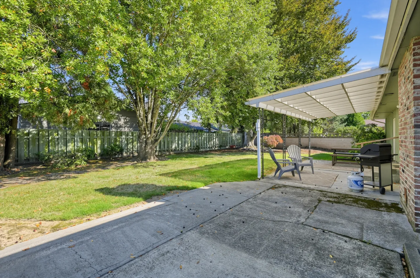 Fenced backyard with a concrete patio, covered awning, barbecue grill, and large shade trees.