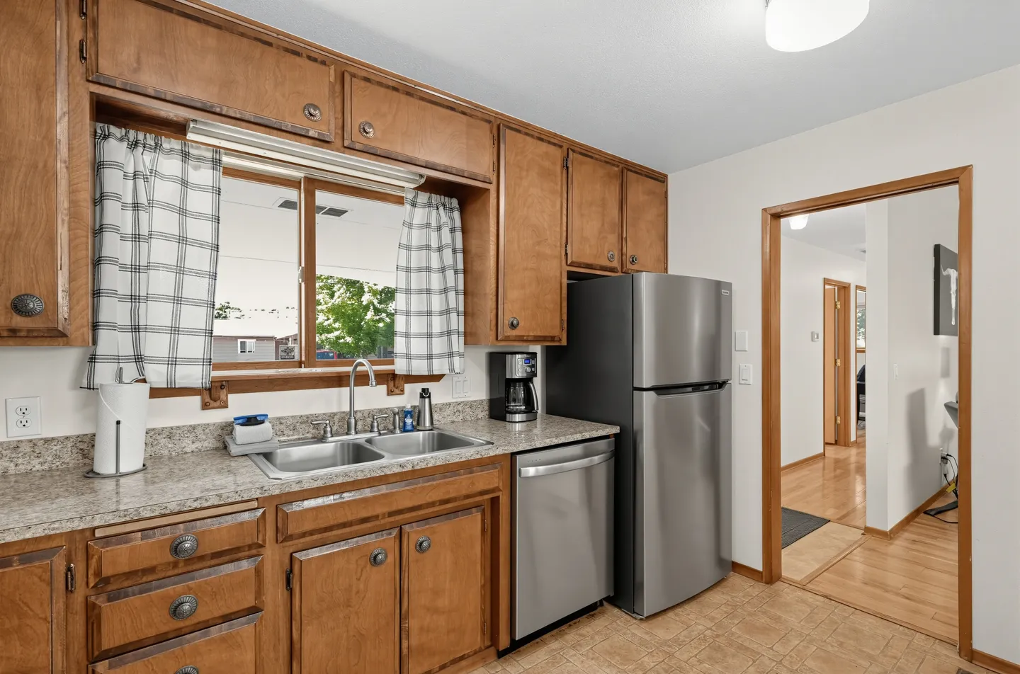 Kitchen featuring wood cabinets, double-basin sink below a window with plaid curtains, and stainless steel appliances.