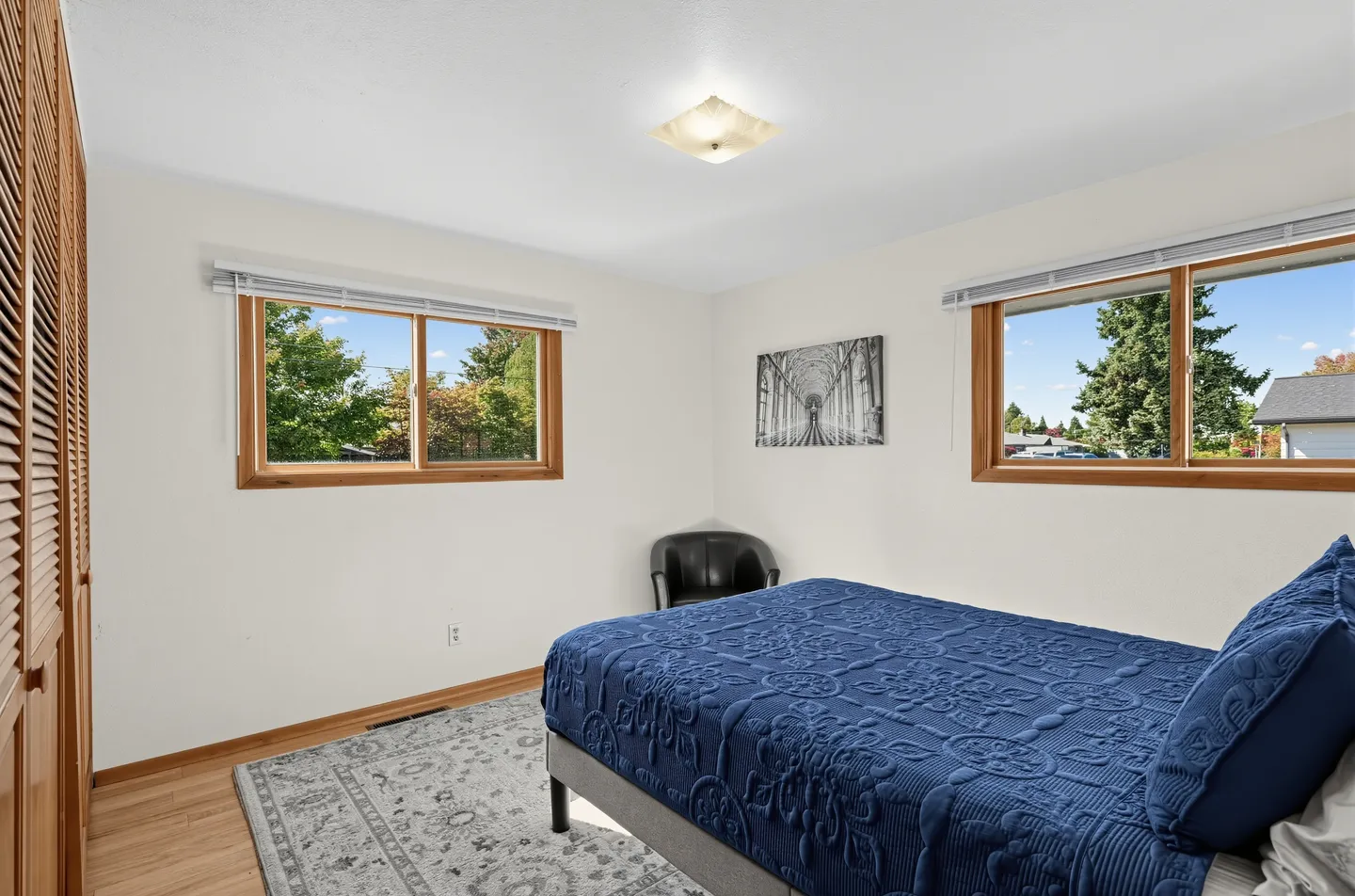 Bedroom with a bed made with a textured blue quilt, a patterned area rug over wood-look flooring, two windows with wood trim, and a corner armchair.