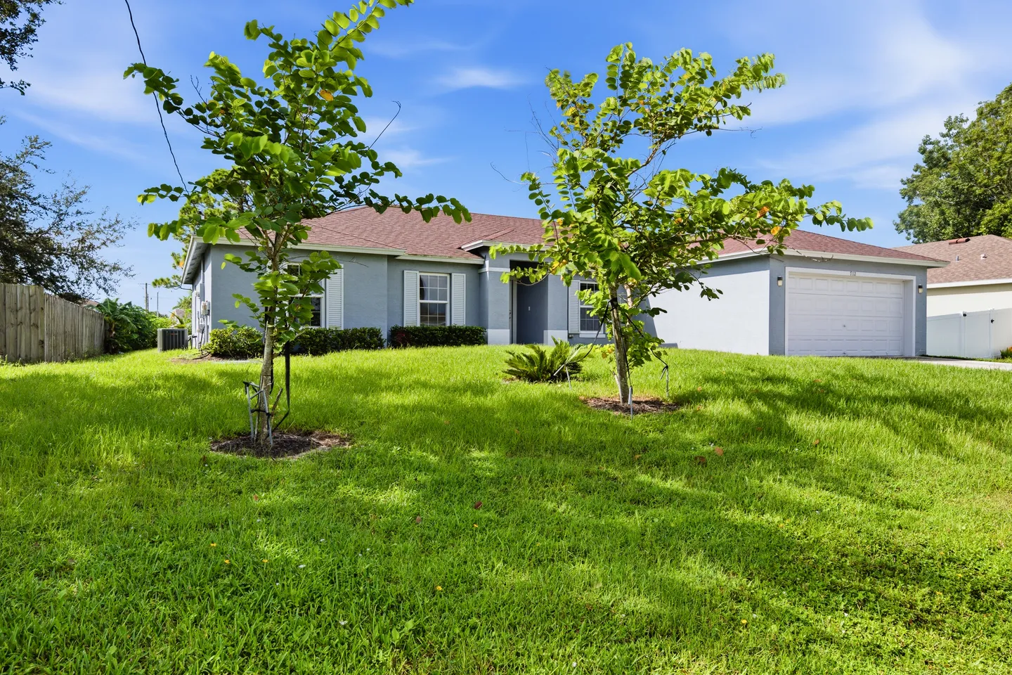 Exterior view of a single-story light blue house with a wide green front lawn, young trees, and an attached two-car garage under a blue sky.