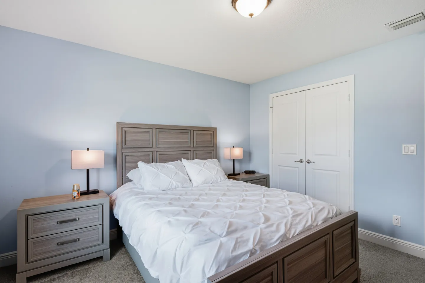 Bedroom with light blue walls, a bed with a wooden paneled headboard, white bedding, two bedside tables with lamps, and a white closet door.