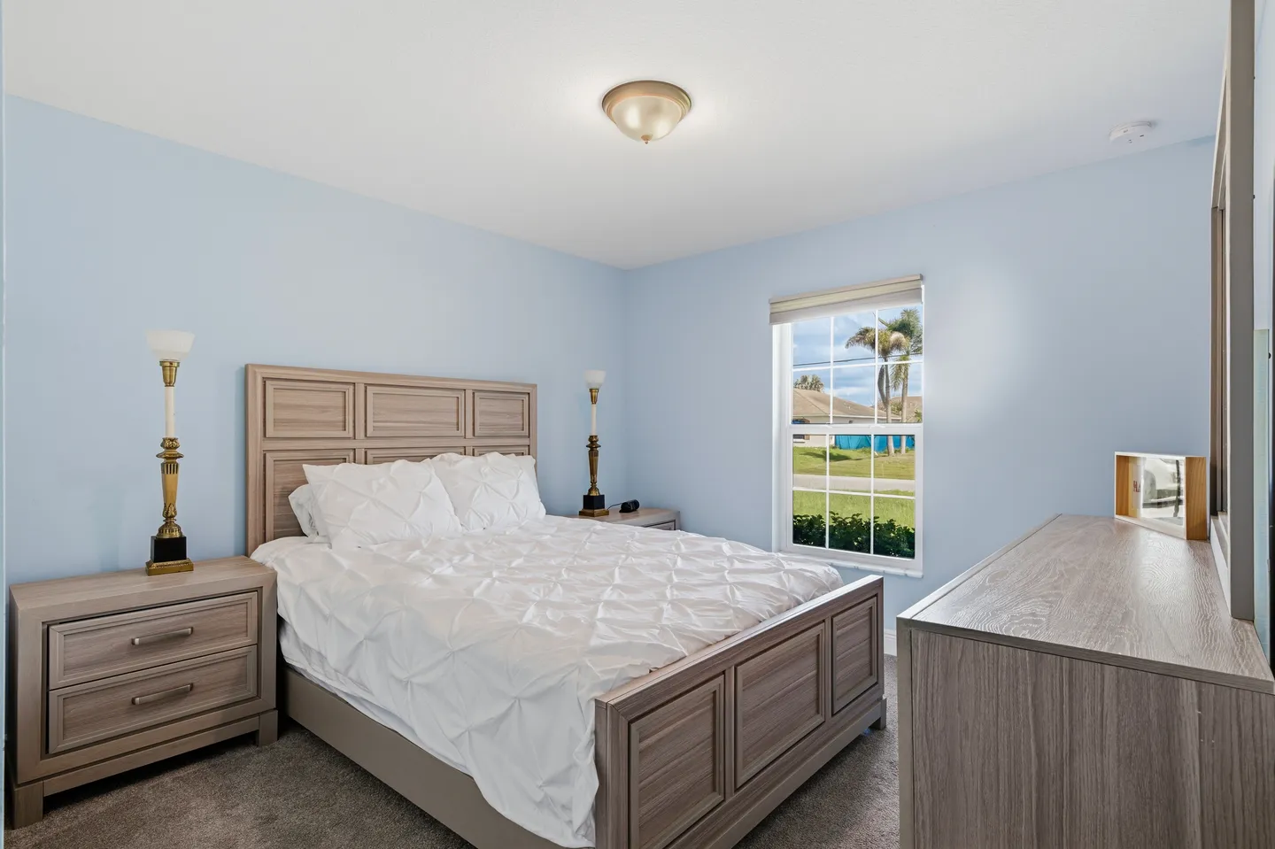 Bedroom with light blue walls, a wooden bed with white bedding, matching nightstands, a dresser, and a window.