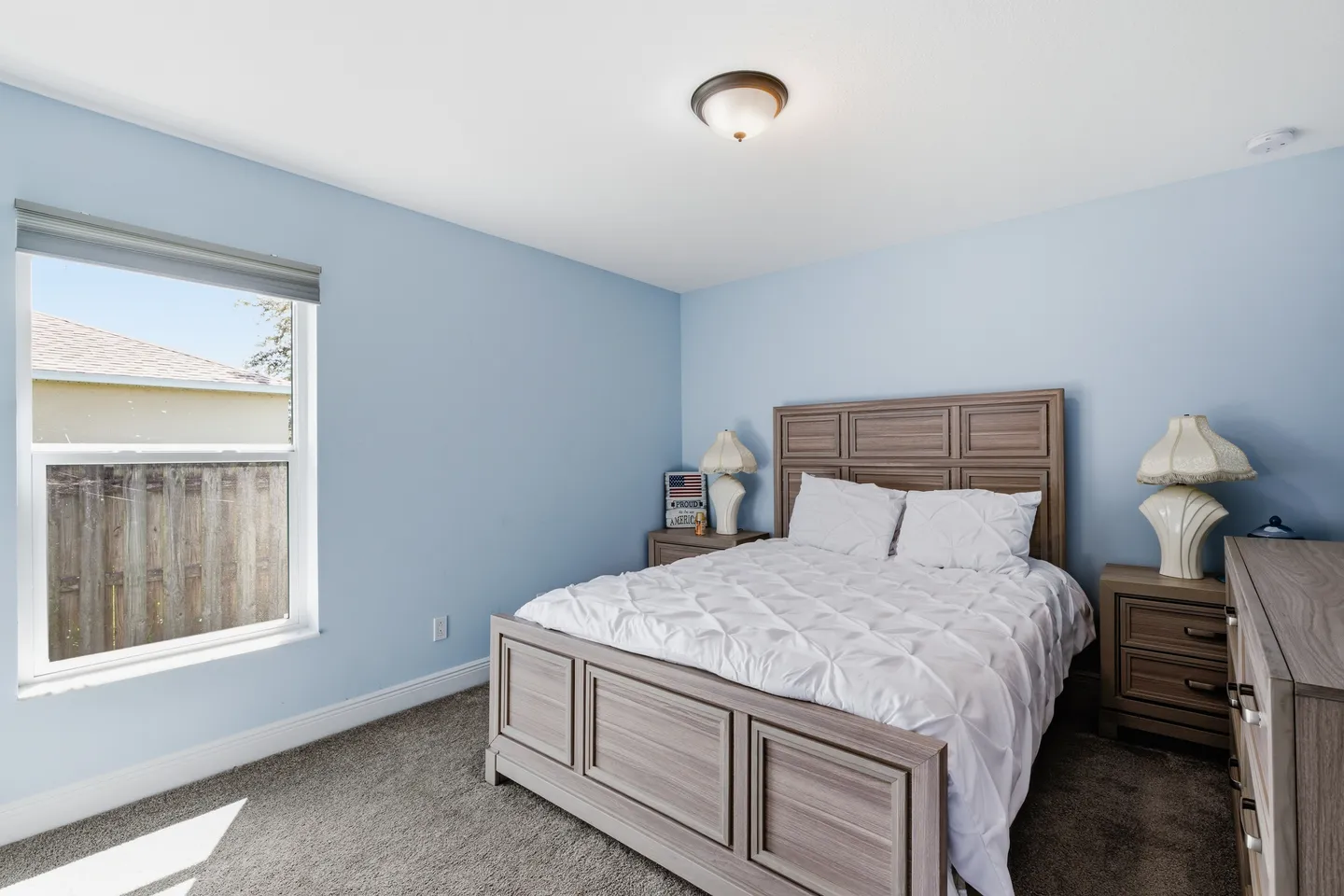 Bedroom with light blue walls, brown patterned carpeting, a wood-paneled bed frame with white bedding, and a window.