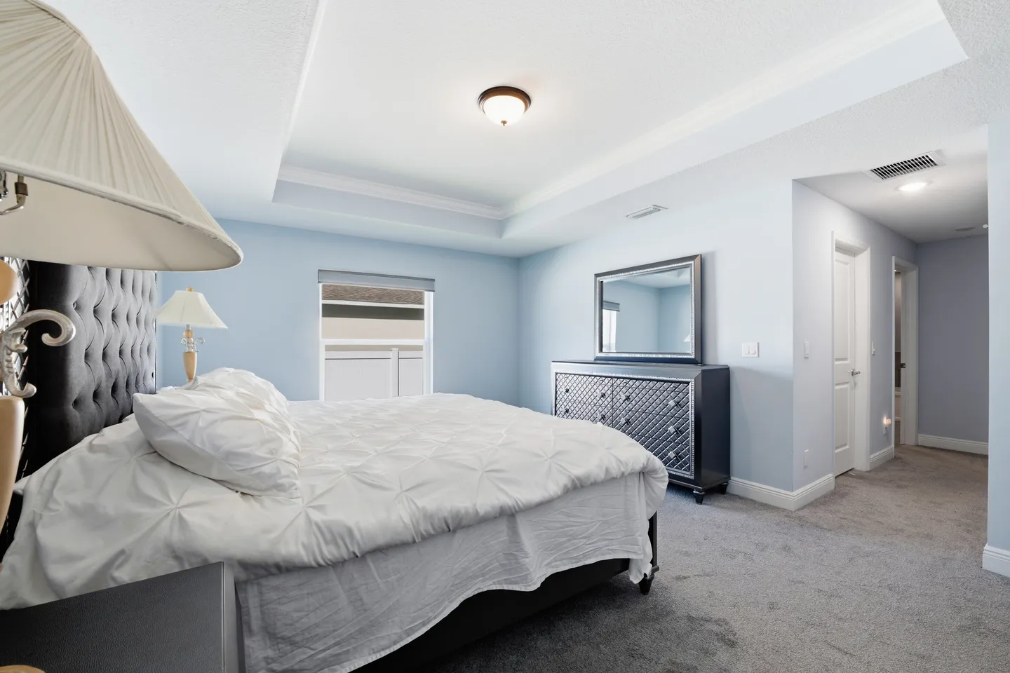Bedroom with light blue walls, tray ceiling, carpeted floor, large bed with tufted headboard, and dark dresser with mirror.