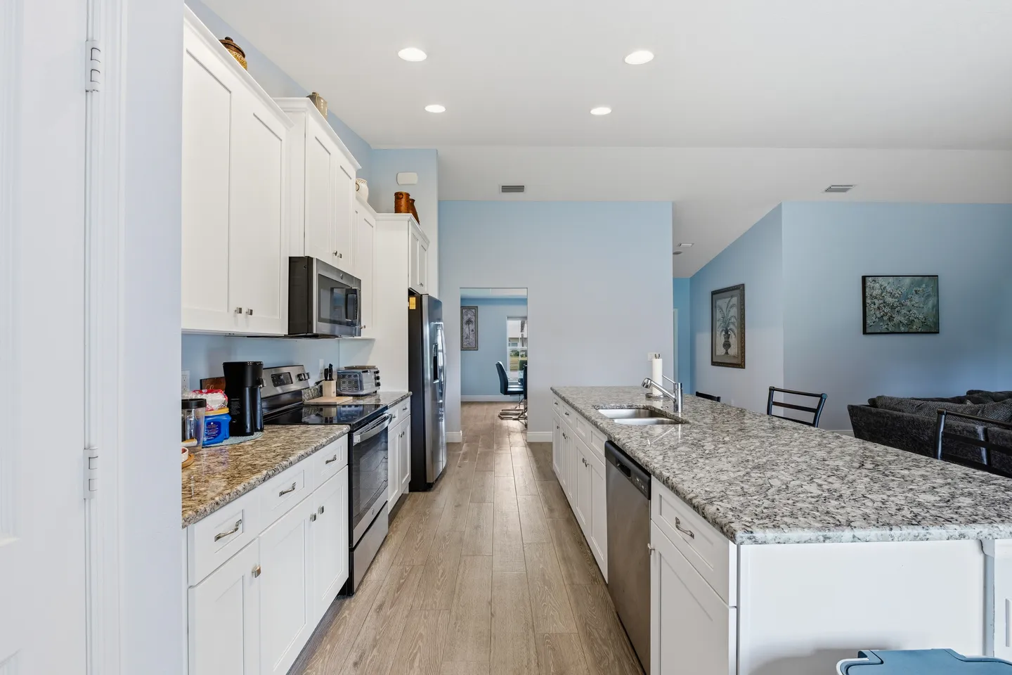 A brightly lit kitchen featuring white shaker cabinets, speckled granite countertops, stainless steel appliances, and a central island with a double sink.