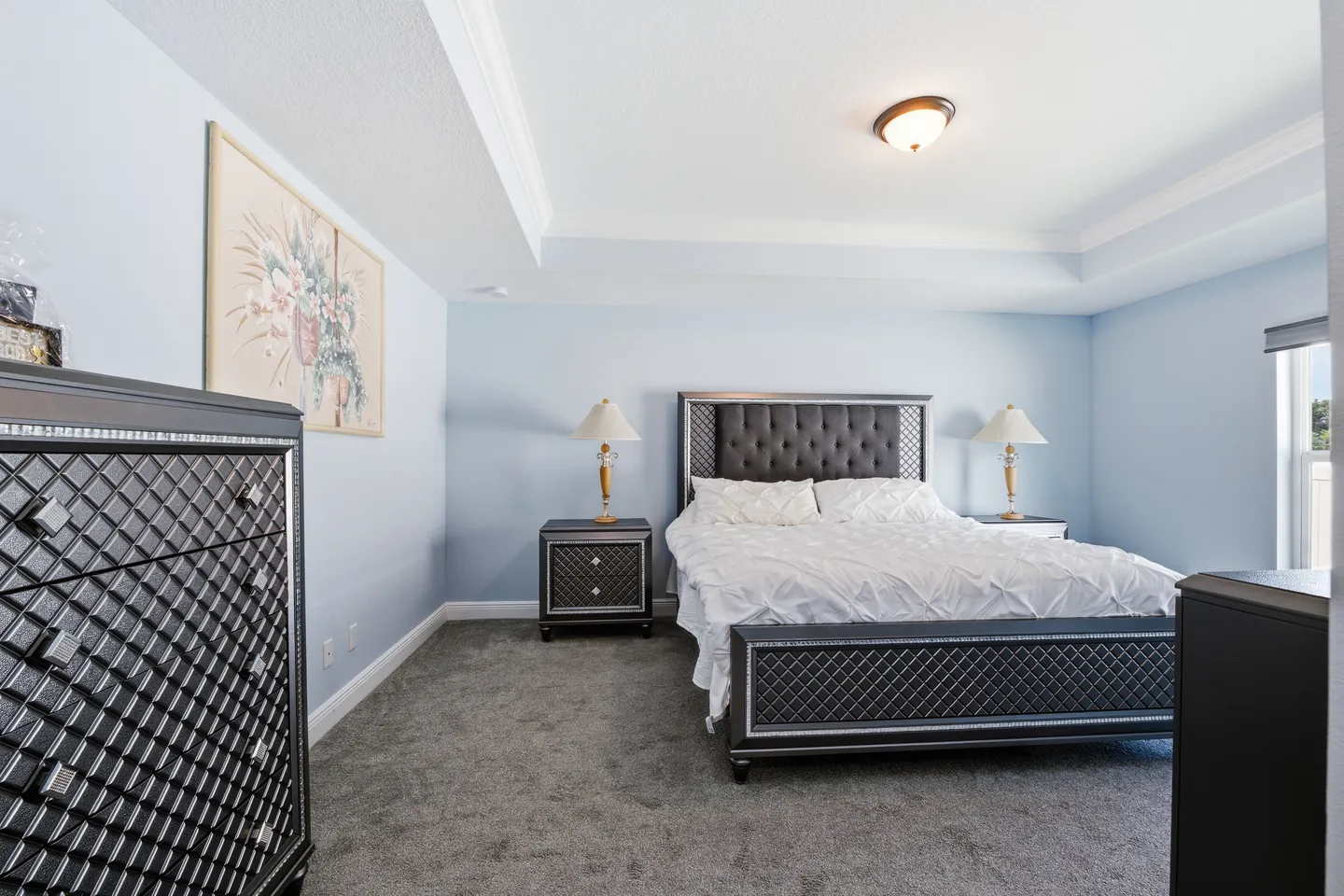 Bedroom with blue walls, tray ceiling, carpeted floor, a bed with a tufted headboard, matching dark furniture, and bedside lamps.