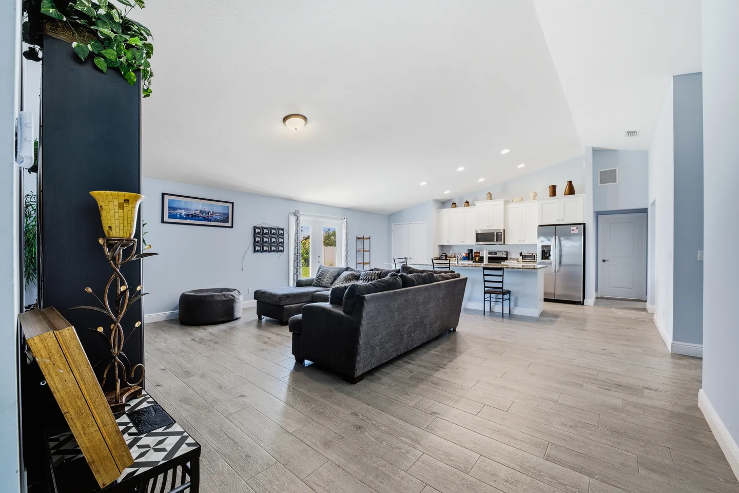 Open-concept living room and kitchen featuring vaulted ceilings, light-colored plank flooring, a dark sectional sofa, and white kitchen cabinets.