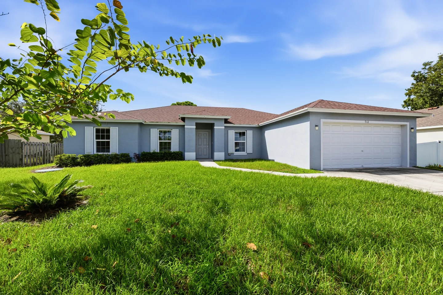 Exterior front view of a single-story light blue house with a two-car garage, manicured lawn, and blue sky.