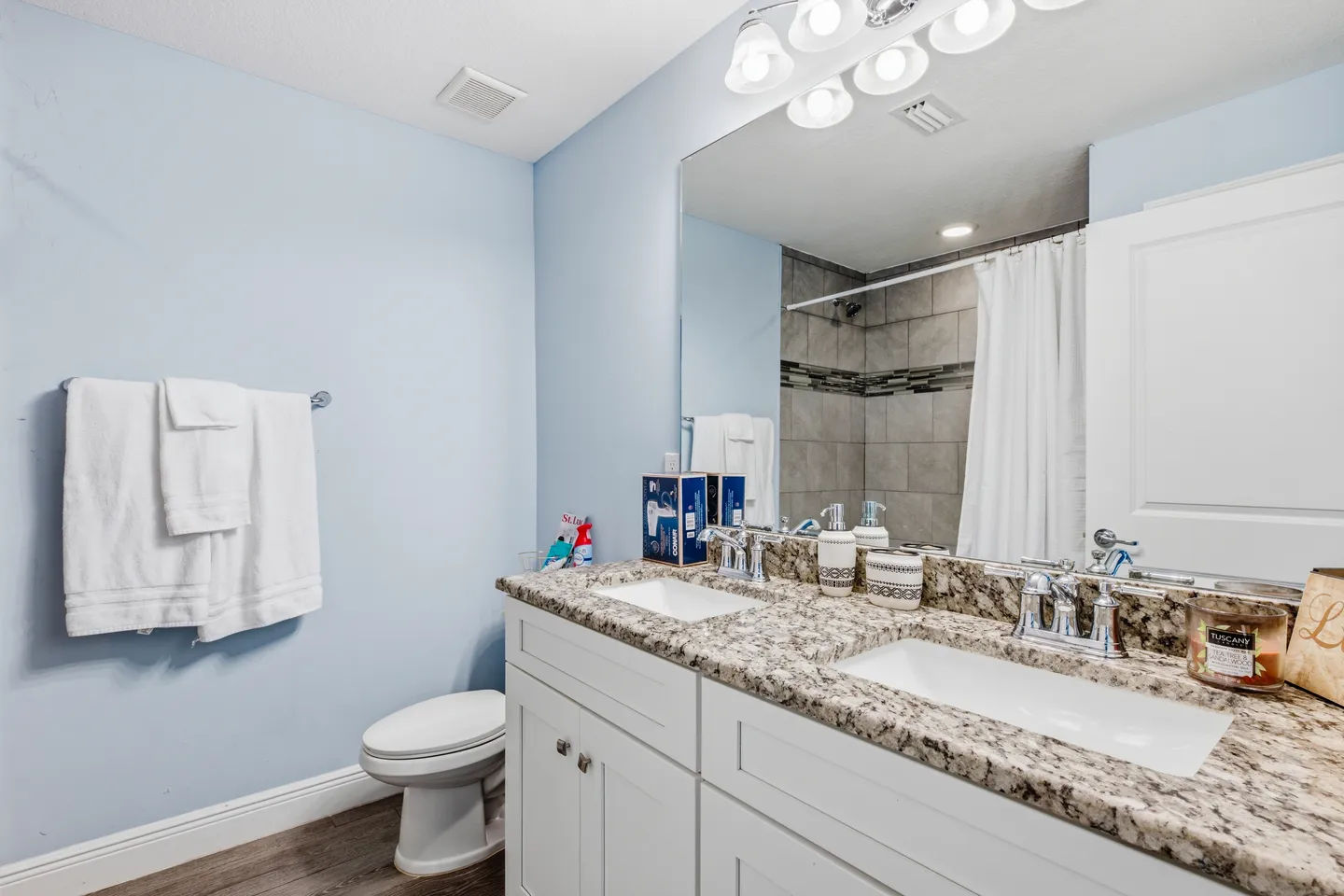Bathroom with a double-sink granite countertop vanity, a large mirror, a toilet, and light blue walls.