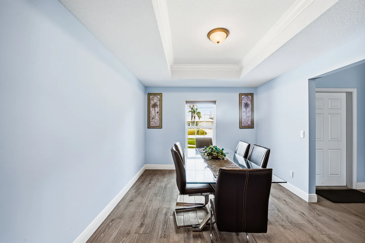 Dining room with light blue walls, wood-look flooring, a glass-top dining table, and dark chairs under a tray ceiling.
