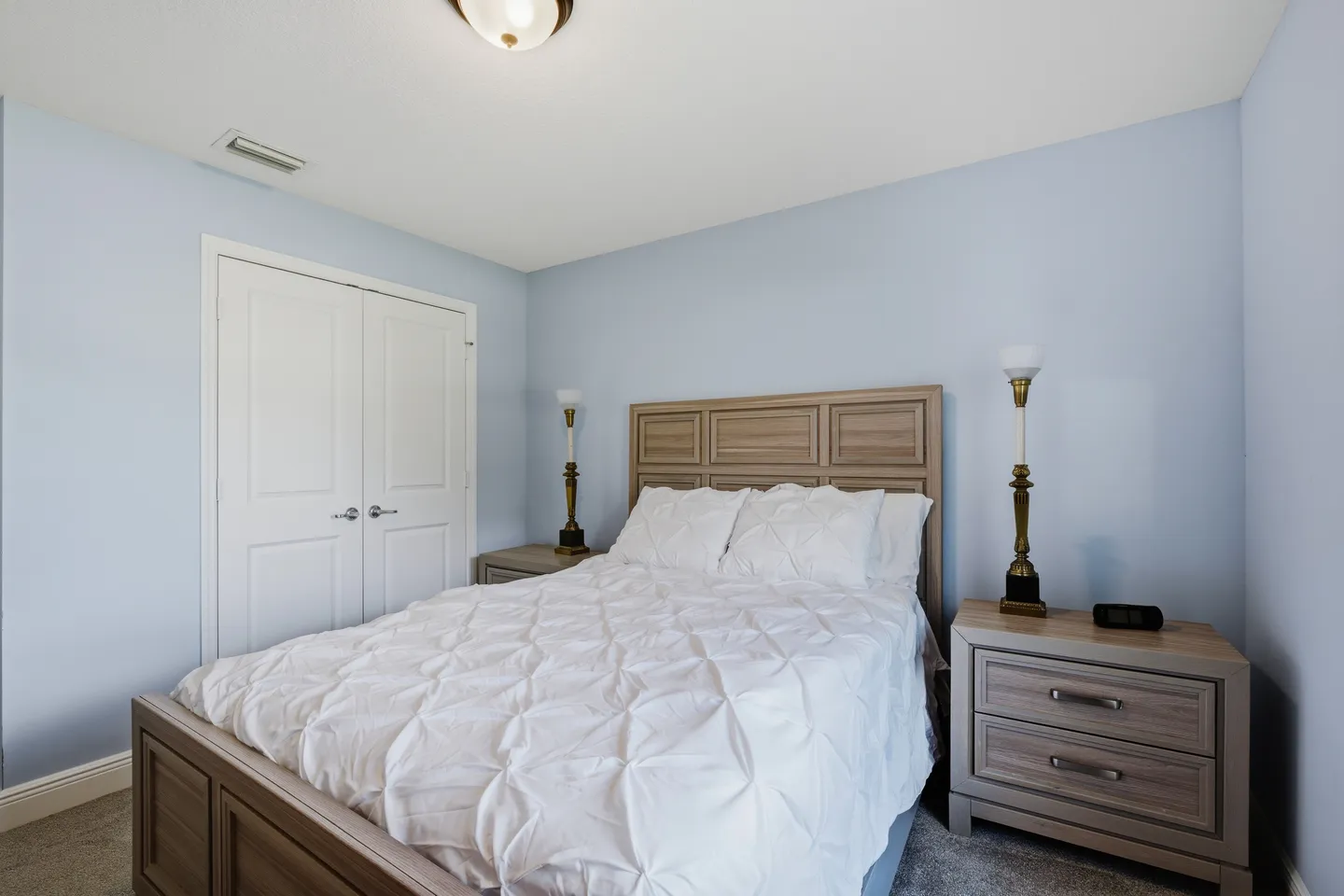 Bedroom with light blue walls, a wooden bed with white bedding, matching nightstands with lamps, and double closet doors.