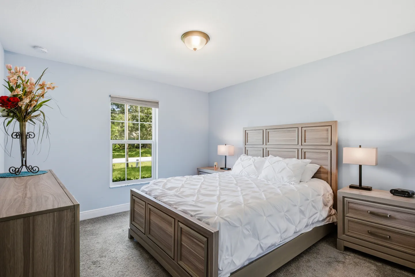 Bedroom with a wooden panel bed, white bedding, matching nightstands with lamps, a dresser, and a window overlooking greenery.