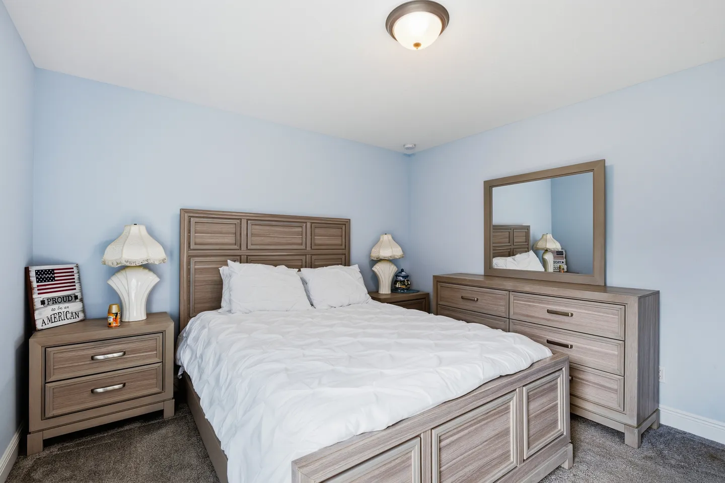 Bedroom with light blue walls, carpet, a wooden paneled bed with white bedding, matching nightstands, and a dresser with a mirror.