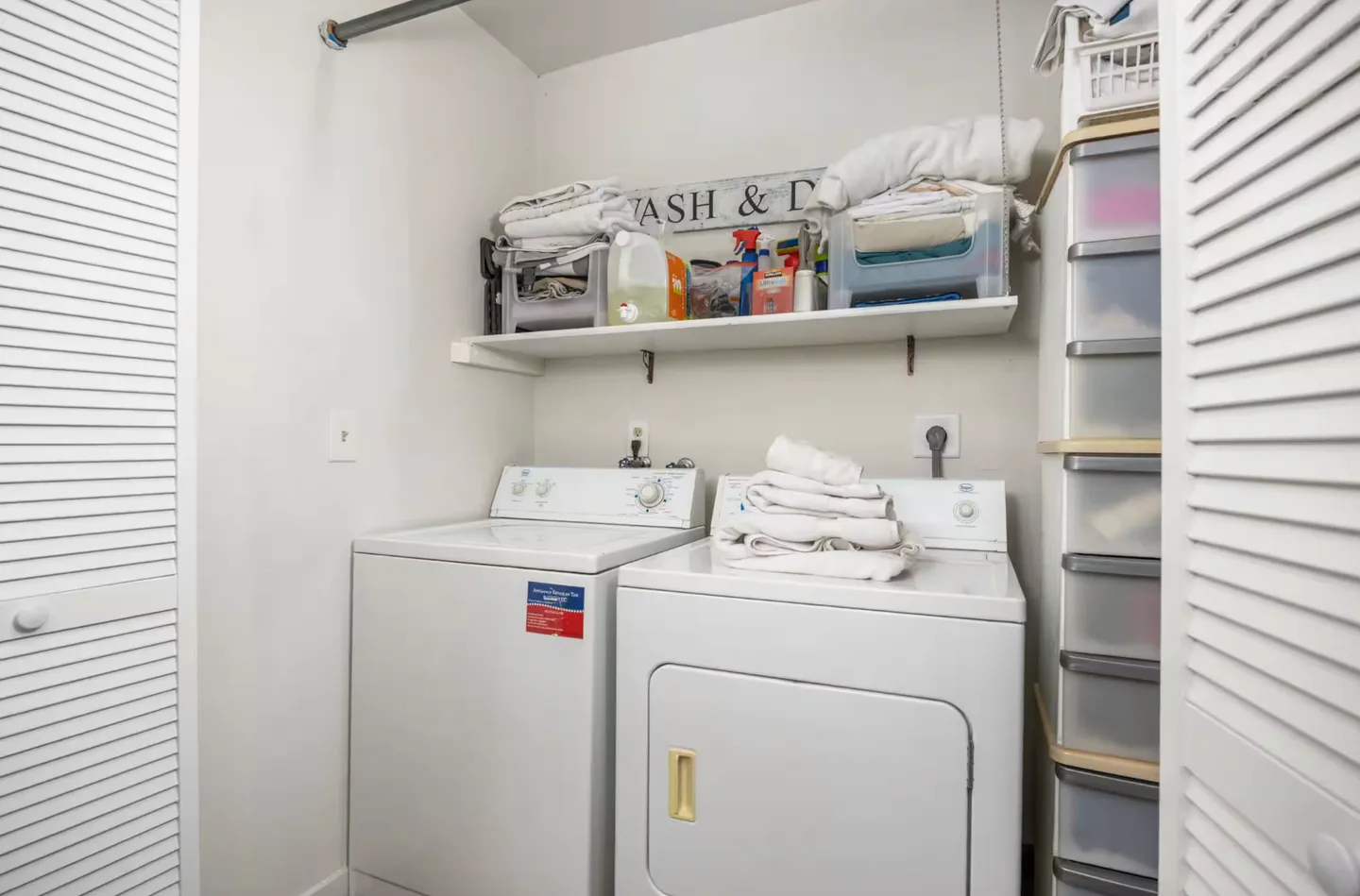 A laundry closet featuring a white washing machine, white dryer, storage shelf, and plastic drawer organizer behind louvered doors.