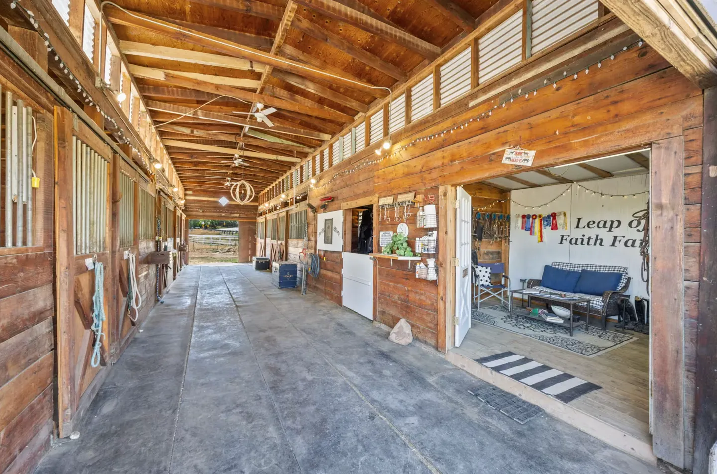 Wide interior breezeway of a wooden horse barn with stalls on both sides and an open tack room or seating area.