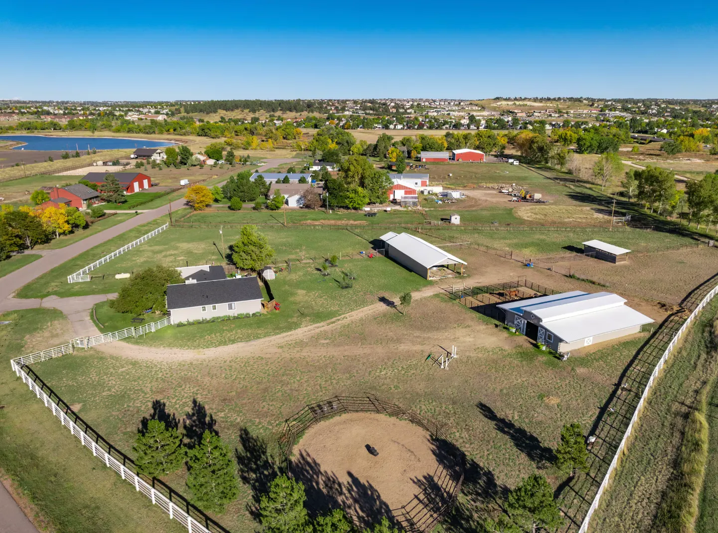 Aerial view of a rural property featuring a house, fenced pastures, barns, and an equestrian round pen under a clear blue sky.