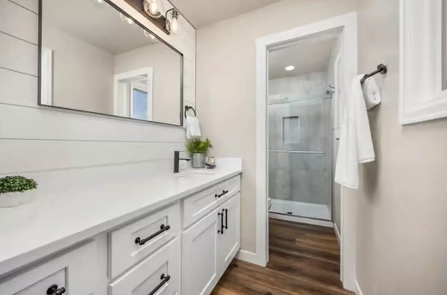 Bathroom with white shiplap accent wall, white vanity with black hardware, mirror, and doorway leading to a walk-in shower.