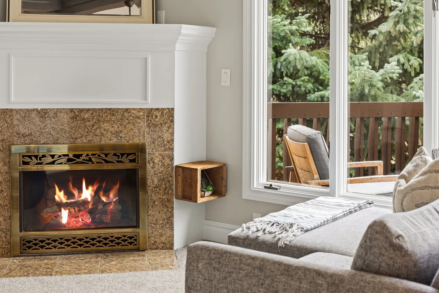 A cozy fireplace corner in a living room next to a large window revealing an outdoor deck and pine trees.