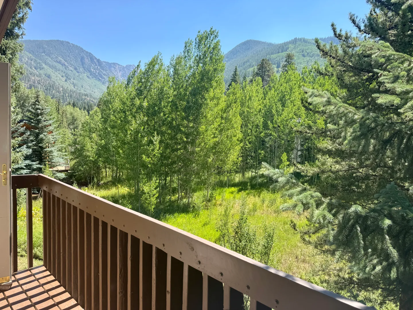 View from an elevated wooden deck looking out over green aspen trees and forested mountains under a clear blue sky.