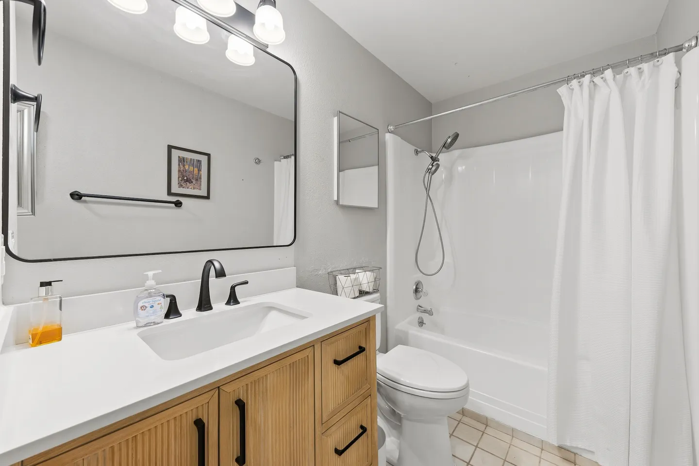 Bathroom with a light wood vanity, white countertop, black fixtures, rounded mirror, toilet, and a white bathtub-shower combo with a white shower curtain.