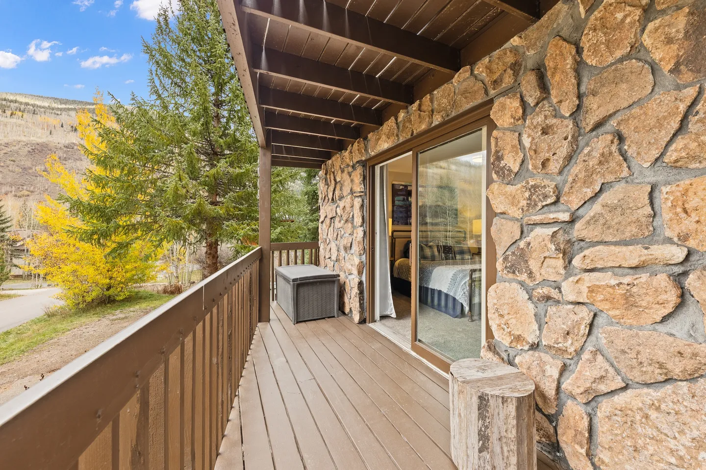 A covered wooden balcony featuring a stone wall, sliding glass doors leading into a bedroom, and views of trees and hills under a blue sky.