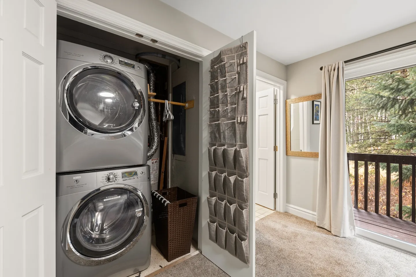 Open laundry closet featuring stacked front-loading washer and dryer next to a room with carpet and sliding glass doors to a deck.