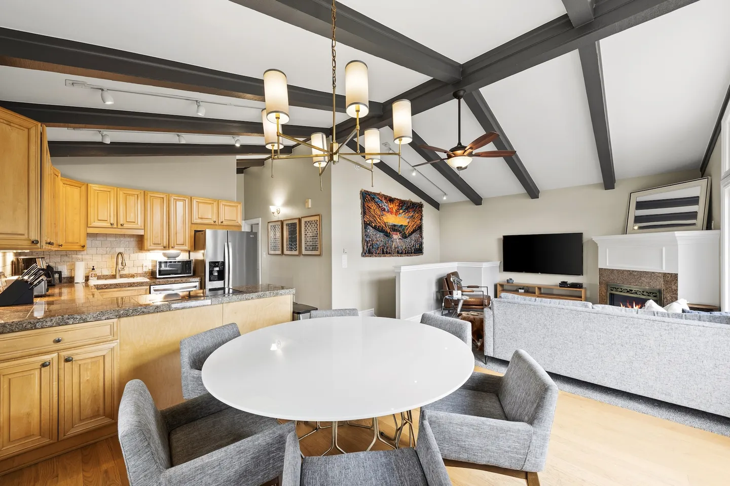 Open-concept dining area with a round white table and grey chairs, looking towards the kitchen and living room with vaulted exposed beam ceiling.