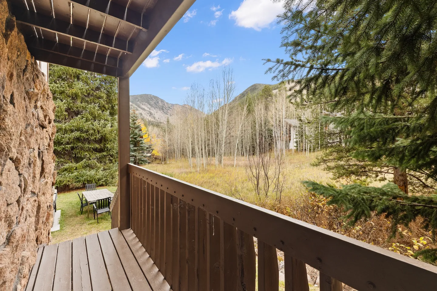 View from an upper-level wooden balcony looking out onto trees, grassy meadow, and distant mountains under a blue sky.