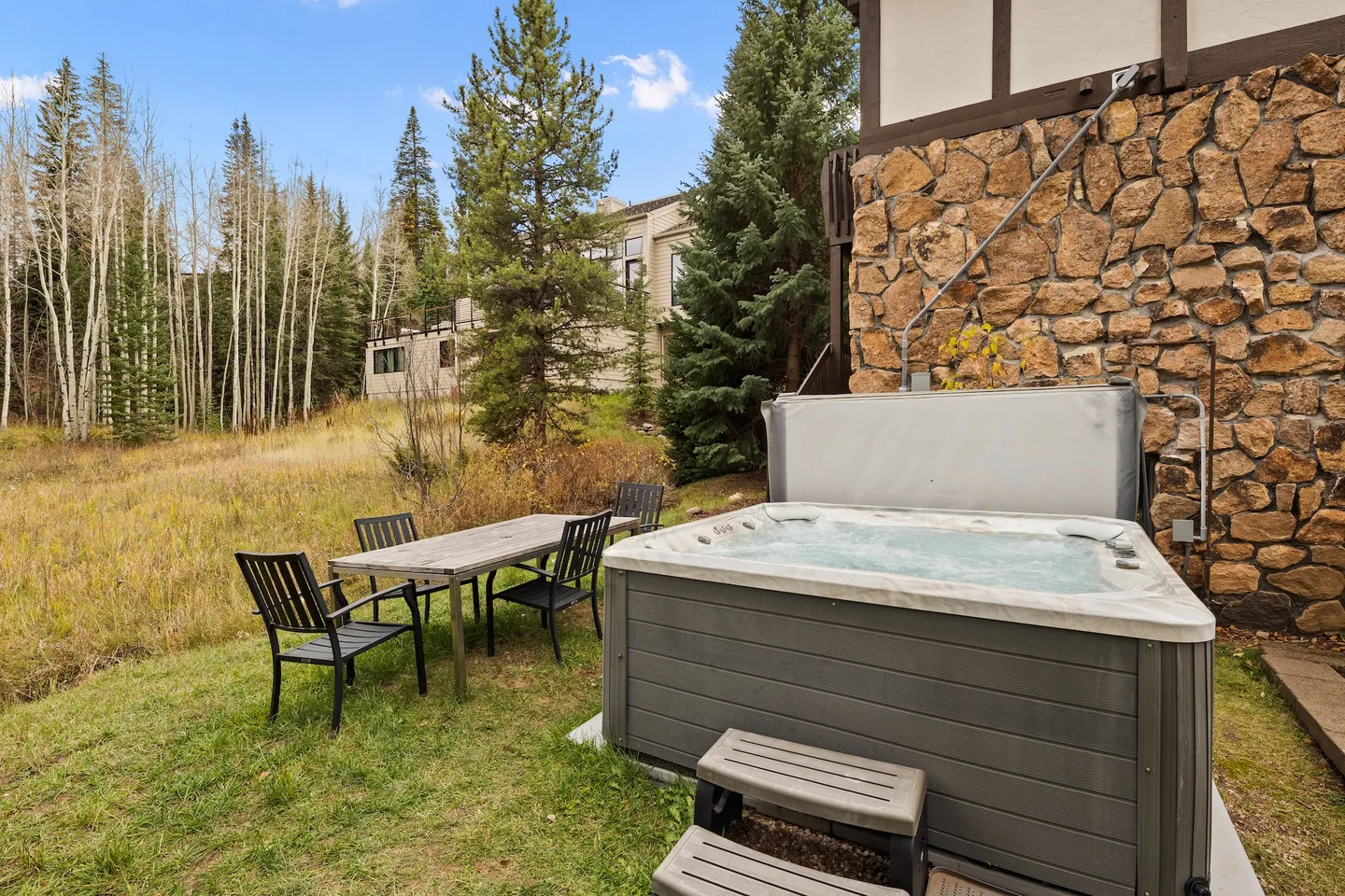 Outdoor hot tub and a dining table with chairs beside a stone wall of a house, facing a grassy meadow and woods.