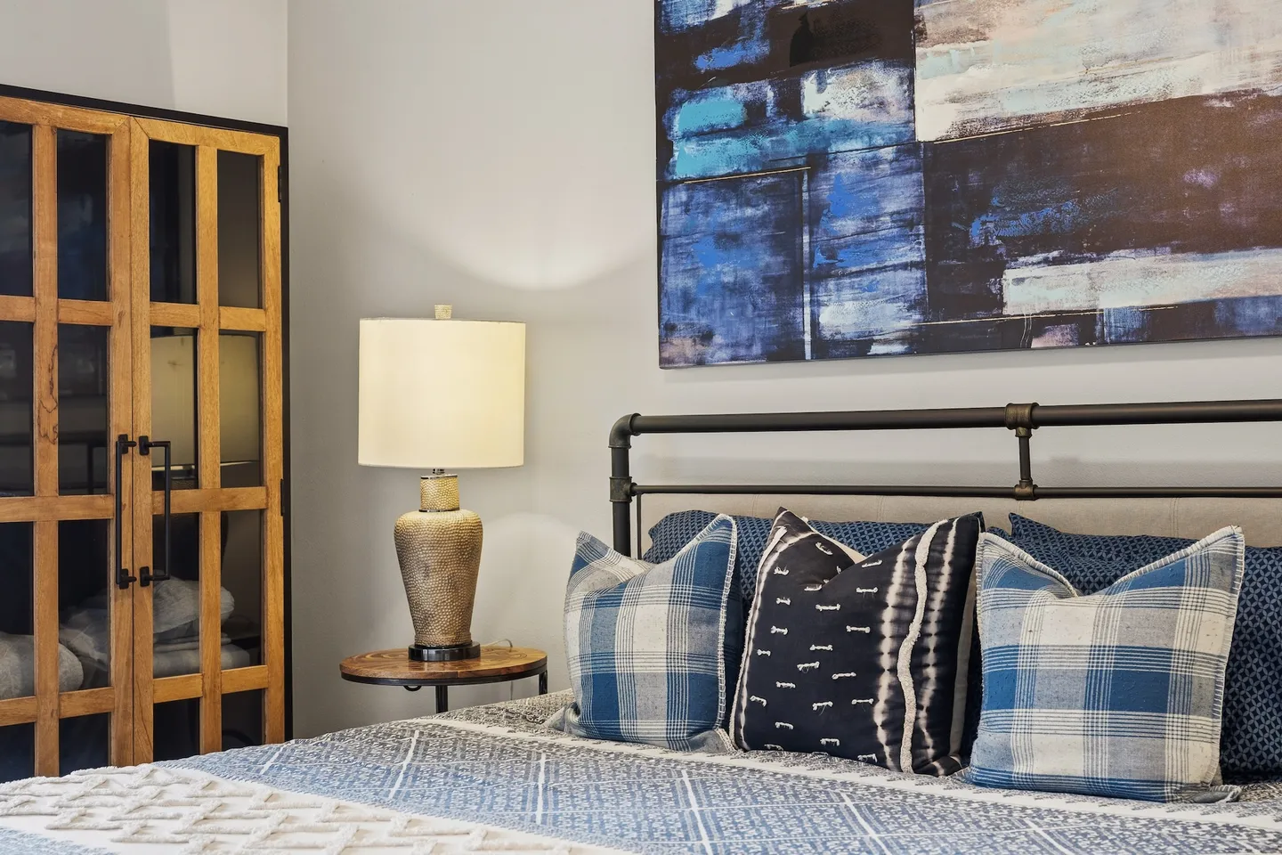 Bedroom detail featuring a dark metal pipe-style headboard with blue plaid and patterned throw pillows, a bedside table with an illuminated lamp, and modern blue wall art.