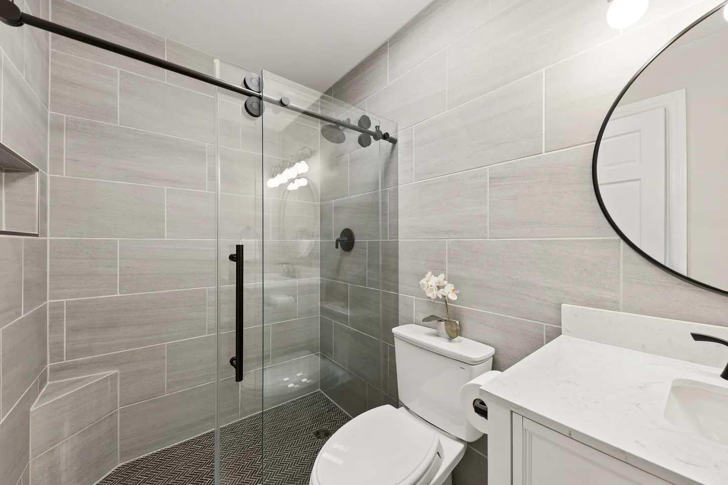 Bathroom with walk-in glass shower, tiled walls, toilet, and vanity with round mirror.