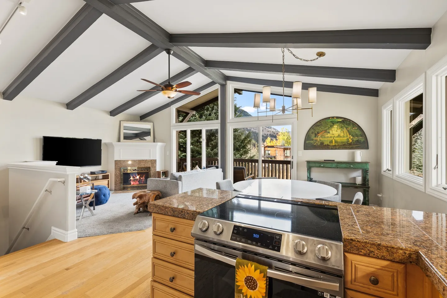 Open-concept living space viewed from a kitchen island, showing exposed ceiling beams, a dining table, a living area with a fireplace, and large windows looking outdoors.