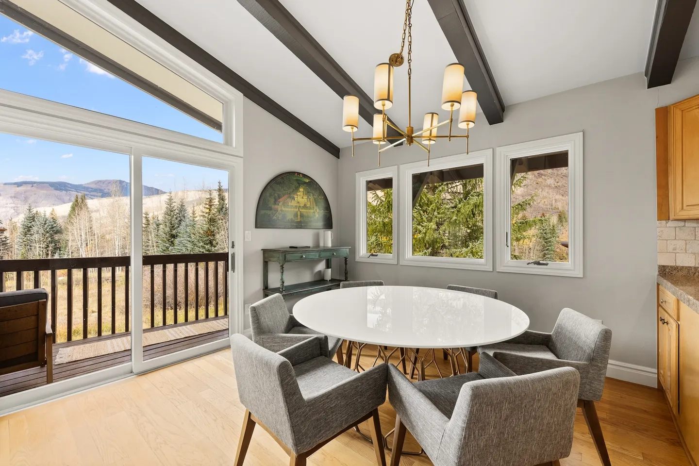 Dining area with a round white table, grey upholstered chairs, modern brass chandelier, and sliding glass doors leading to an outdoor deck with mountain views.