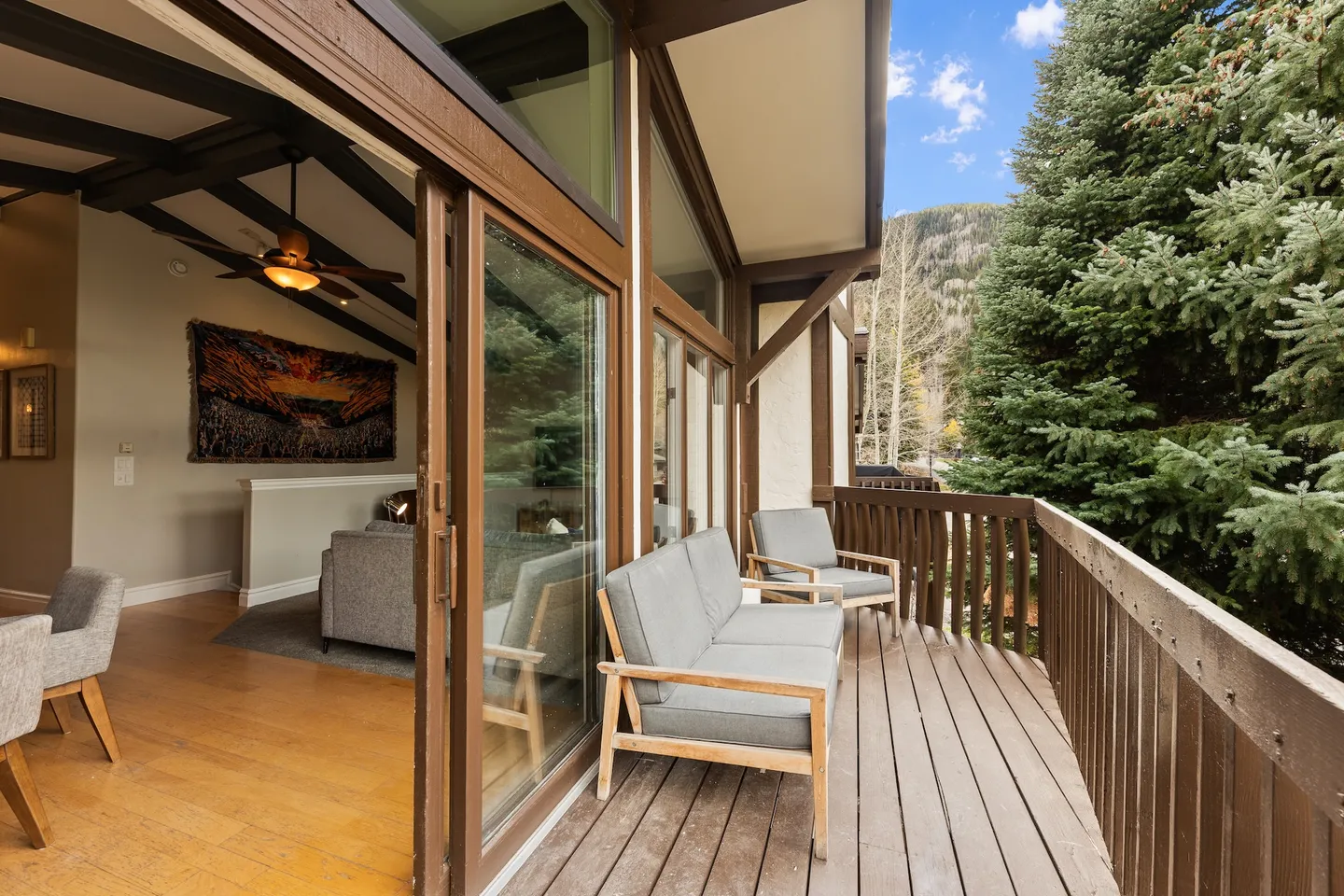 Balcony with wooden decking and outdoor seating overlooking evergreen trees, connected via sliding glass doors to the living room.